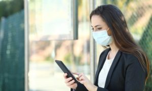 Woman wearing a protective mask using mobile phone sitting in a bus stop